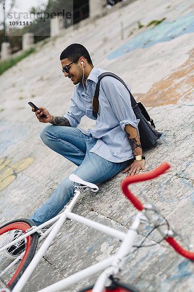 Junger Mann mit einem Fahrrad auf einer Wand sitzend und auf das Handy schauend