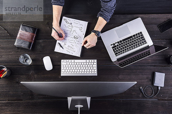Mann arbeitet am Schreibtisch mit Computer und Laptop und macht sich Notizen auf einem Blatt Papier.