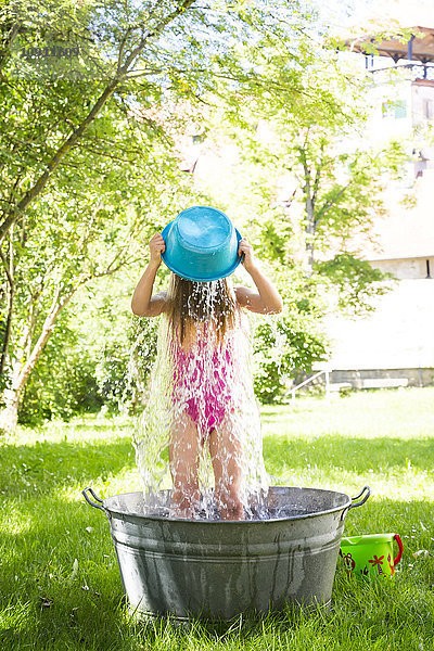 Kleines Mädchen steht in einer Wanne auf einer Wiese und gießt Wasser über sich selbst.