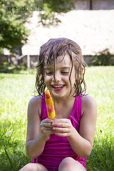 Porträt eines glücklichen kleinen Mädchens mit nassen Haaren  das auf einer Wiese mit Eis am Stiel sitzt.