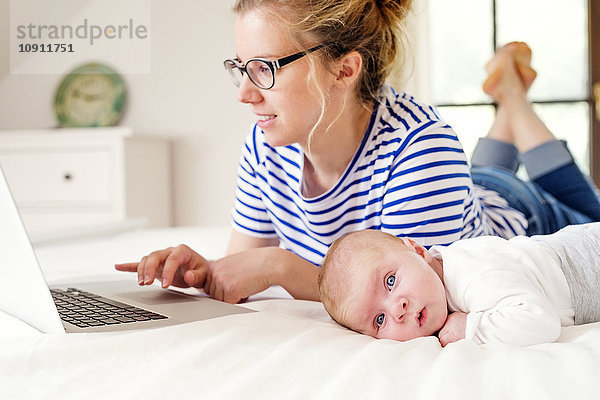 Mutter mit Baby auf dem Bett liegend  mit Laptop arbeitend