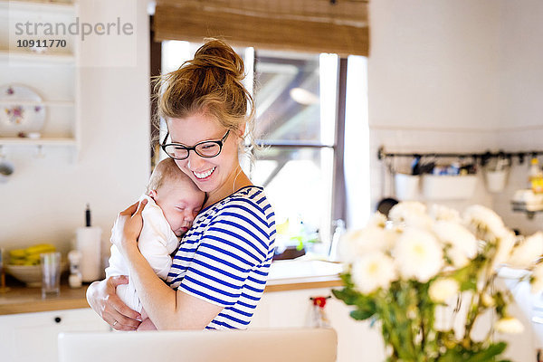 Glückliche Mutter mit Baby in der Küche mit Blick auf Laptop