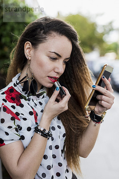 UK  London  junge Frau beim Auftragen von Lippenstift mit Hilfe ihres Smartphones