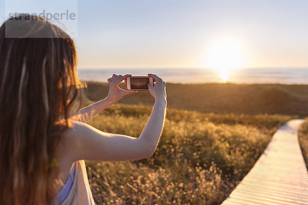 Junge Frau fotografiert Strand bei Sonnenuntergang mit Smartphone