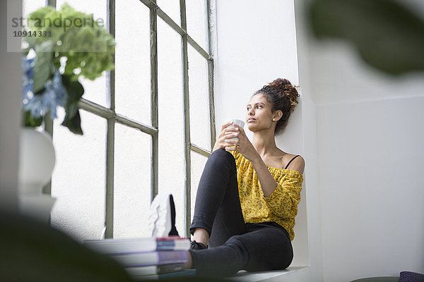 Junge Frau sitzt auf der Fensterbank mit einer Tasse Kaffee
