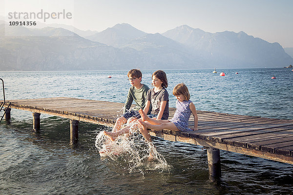 Italien  Brenzone  drei Kinder sitzen Seite an Seite auf dem Steg und spritzen mit Wasser.