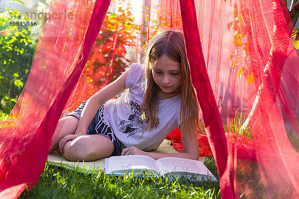 Mädchen sitzt auf einer Wiese unter einem Moskitonetz und liest ein Buch.