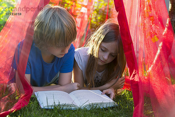 Schwester und Bruder liegen auf einer Wiese unter einem Moskitonetz und lesen zusammen ein Buch.