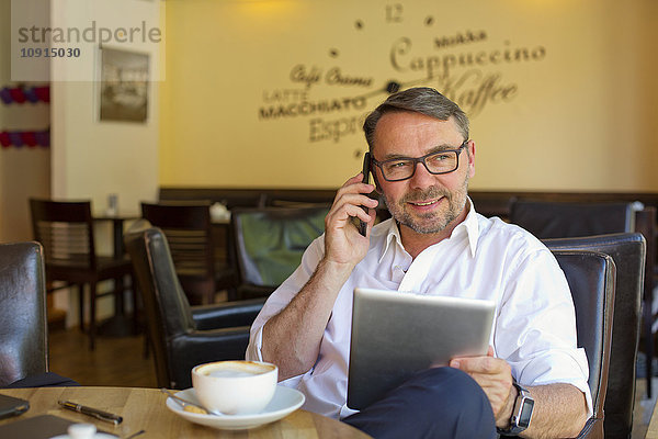 Porträt eines lächelnden Geschäftsmannes mit digitalem Tablett im Café beim Telefonieren mit Smartphone