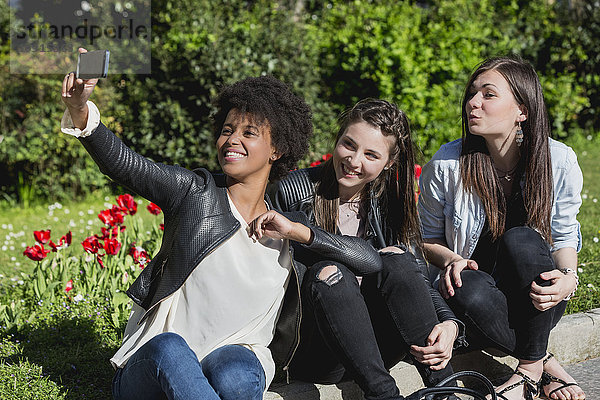 Drei Freundinnen sitzen auf Rasen und nehmen Selfie mit dem Smartphone.