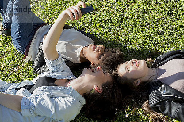 Drei Freundinnen liegen auf Rasen und nehmen Selfie mit dem Smartphone.