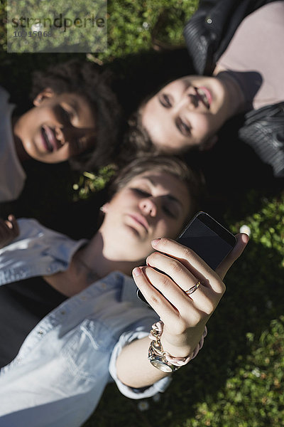 Drei Freundinnen liegen auf Rasen und nehmen Selfie mit dem Smartphone.