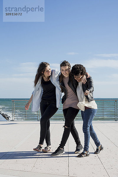 Drei junge Frauen gehen Arm in Arm am Meer.