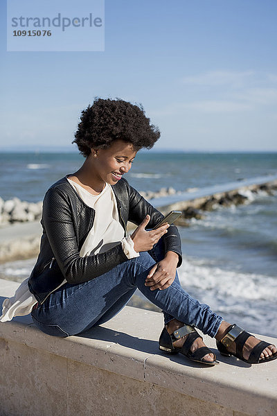 Junge Frau sitzt am Meer und benutzt ein Smartphone.