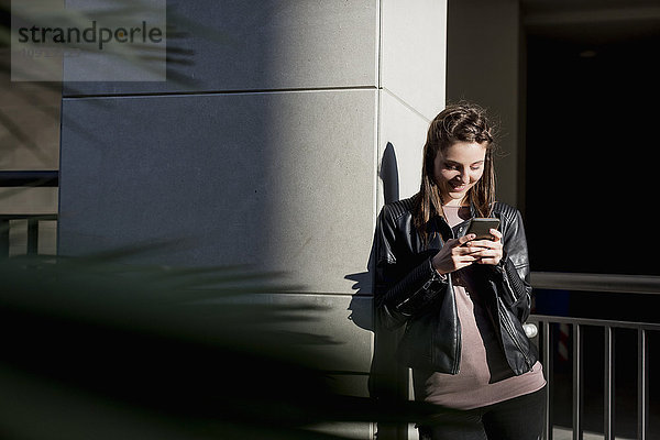 Junge Frau  die sich mit dem Smartphone an die Wand lehnt