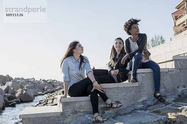 Drei junge Frauen sitzen an der Wand am Meer.
