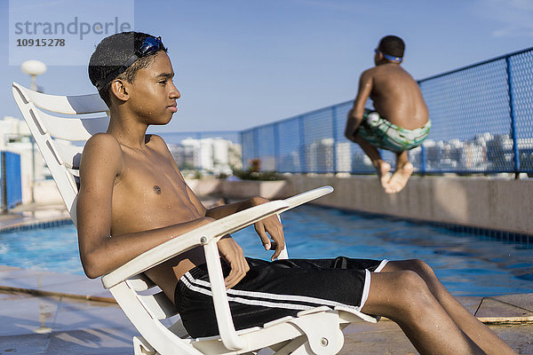 Teenager-Junge sitzt auf einem Stuhl vor dem Schwimmbad  während sein Freund einen Kanonenkugel-Tauchgang macht.