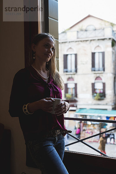 Junge Frau steht am Fenster und trinkt Kaffee.