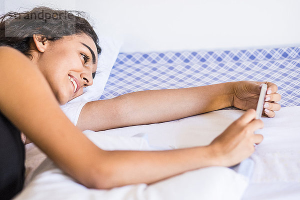Lächelndes Teenagermädchen liegt auf dem Bett und schaut auf das Smartphone.