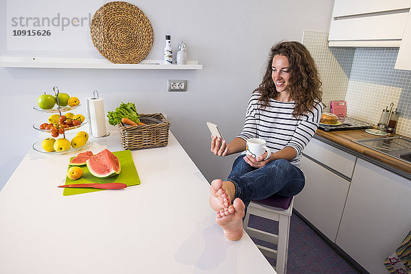 Lächelnde Frau sitzt mit erhobenen Füßen in der Küche und schaut auf das Handy.