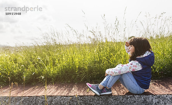 Kleines Mädchen sitzt auf dem Bürgersteig vor einer Wiese und schaut in die Ferne.
