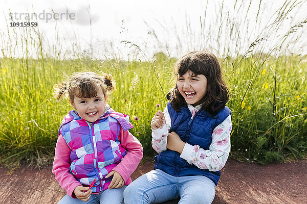 Zwei lachende kleine Schwestern sitzen vor einer Wiese.