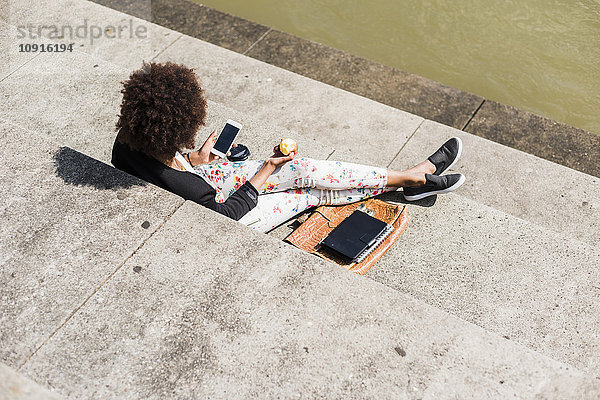 Junge Frau sitzt auf einer Treppe und isst einen Apfel  während sie auf ihr Smartphone schaut.