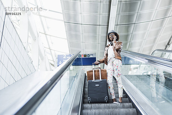 Junge Frau mit Gepäck auf Rolltreppe stehend telefonierend mit Smartphone
