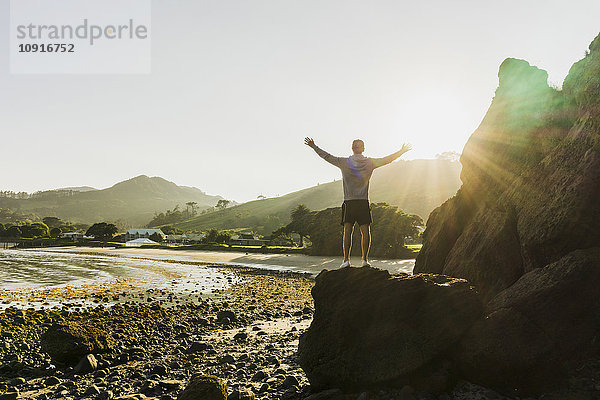 Neuseeland  Whangamata  Rückansicht eines glücklichen Mannes auf einem Felsen mit Blick auf den Strand
