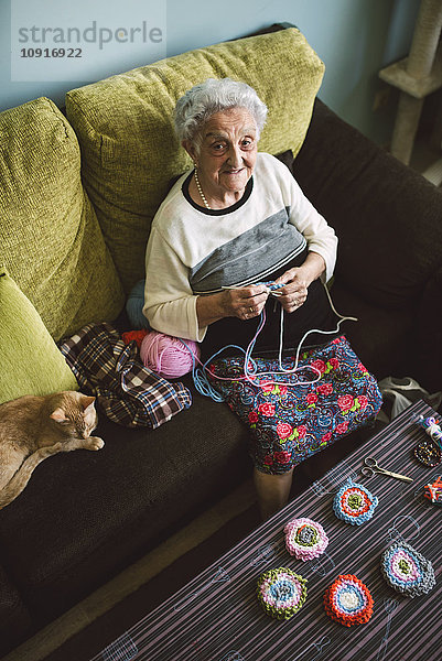 Porträt der häkelnden Seniorin  die neben ihrer schlafenden Katze auf der Couch sitzt.