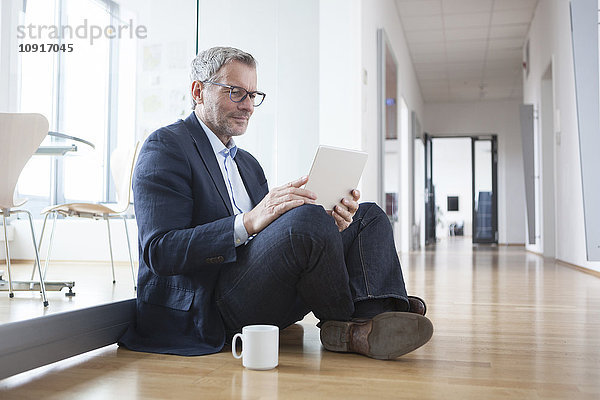 Erfolgreicher Geschäftsmann sitzt auf dem Boden und benutzt ein digitales Tablett in seinem Büro.