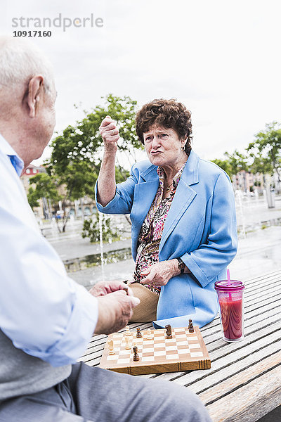 Porträt einer älteren Frau  die auf einer Bank sitzt und mit ihrem Mann Schach spielt.