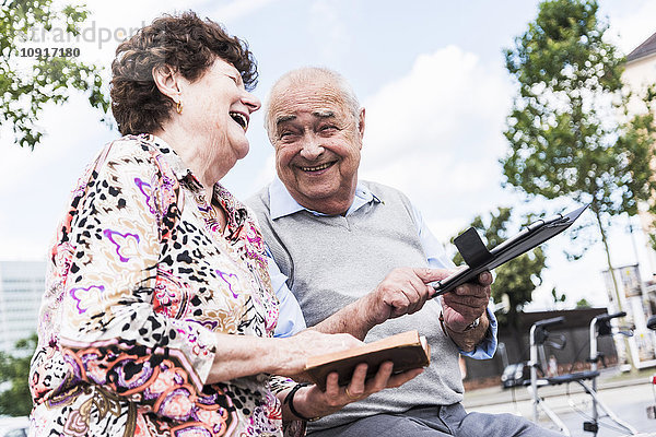 Lachendes Seniorenpaar sitzend auf einer Bank mit Buch und Tablette