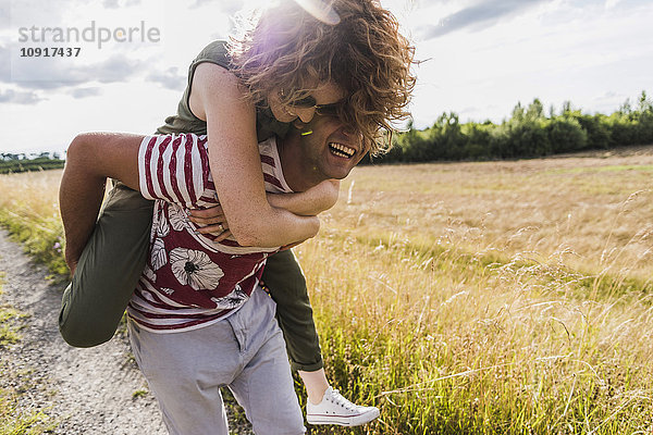 Glücklicher junger Mann trägt Freundin Huckepack auf Feldweg