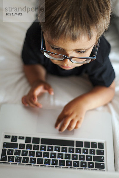 Kleiner Junge mit Brille auf dem Bett liegend mit Laptop