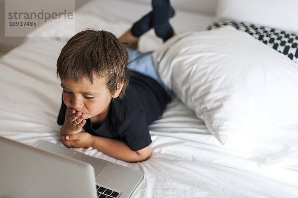 Lächelnder kleiner Junge auf dem Bett liegend mit Laptop