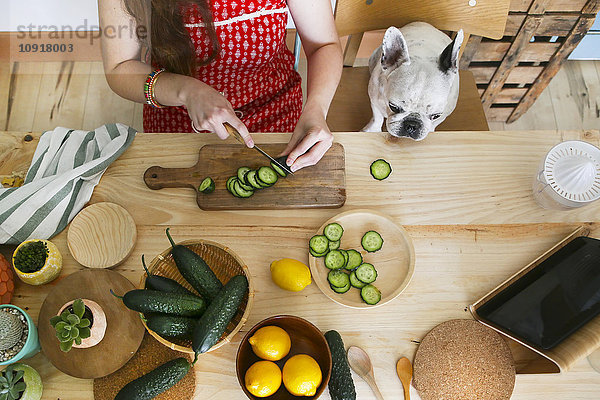 Französische Bulldogge beobachtet Frau beim Gurkenschneiden auf dem Tisch
