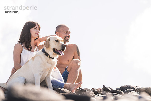 Paar sitzend mit ihrem Hund am steinigen Strand