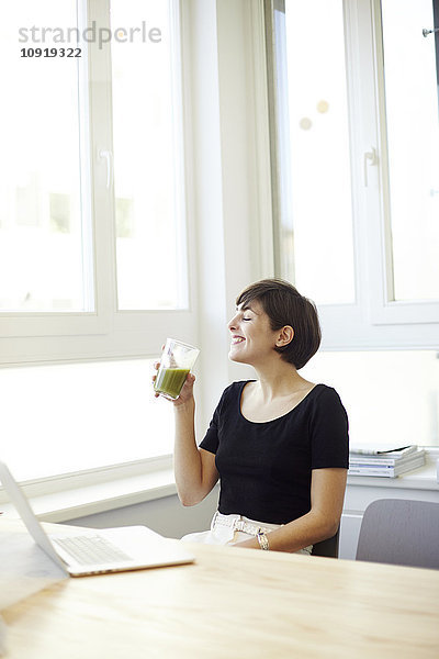 Glückliche Frau trinkt grünen Smoothie im Büro