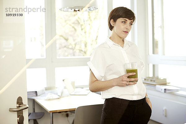 Tagträumende Frau mit einem Glas grünen Smoothie im Büro