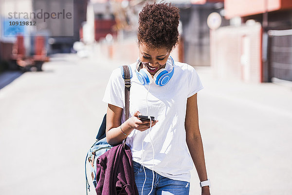Lächelnde junge Frau mit Kopfhörer und Blick auf das Handy im Freien