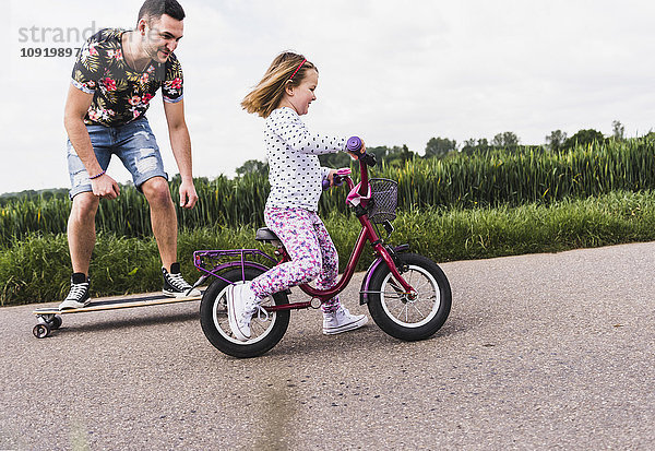 Vater auf dem Skateboard begleitet Tochter auf dem Fahrrad