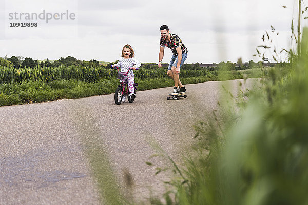 Vater auf dem Skateboard begleitet Tochter auf dem Fahrrad