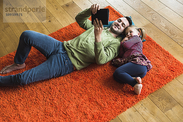 Vater und Tochter liegen auf einem Teppich auf dem Boden mit Hilfe eines digitalen Tabletts.