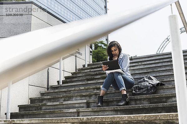 Junge Frau sitzt auf einer Treppe und schaut auf ein digitales Tablett.