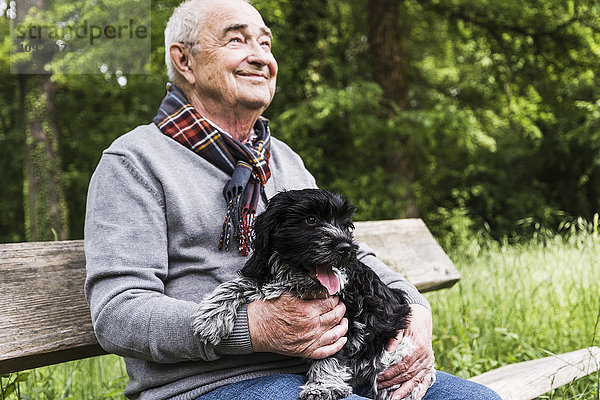 Lächelnder älterer Mann sitzt mit seinem Hund auf einer Bank in der Natur.