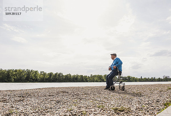 Älterer Mann sitzt auf einem Rollator am Flussufer.