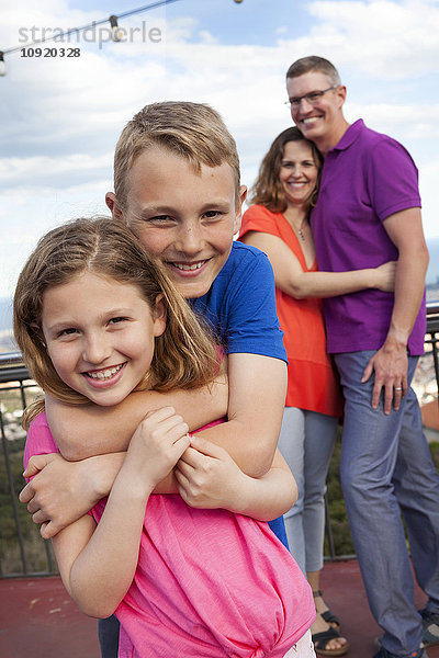 Porträt eines Jungen  der seine kleine Schwester umarmt  während seine Eltern im Hintergrund stehen.