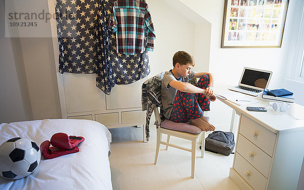 Gelangweilter Junge macht Hausaufgaben am Schreibtisch im Schlafzimmer