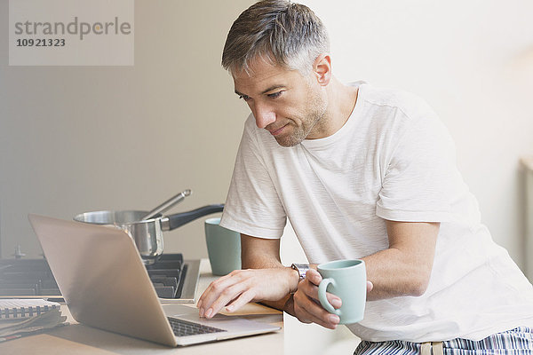 Mann im Pyjama  der Kaffee trinkt und am Laptop in der Küche arbeitet.